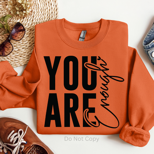 An orange sweatshirt with the text 'you are enough' printed in black on the front, placed on a table with sunglasses, a hat, and shoes around it.