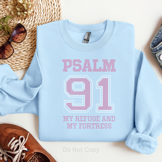 Light blue sweatshirt with 'PSALM 91 MY REFUGE AND MY FORTRESS' text on a white surface with accessories.