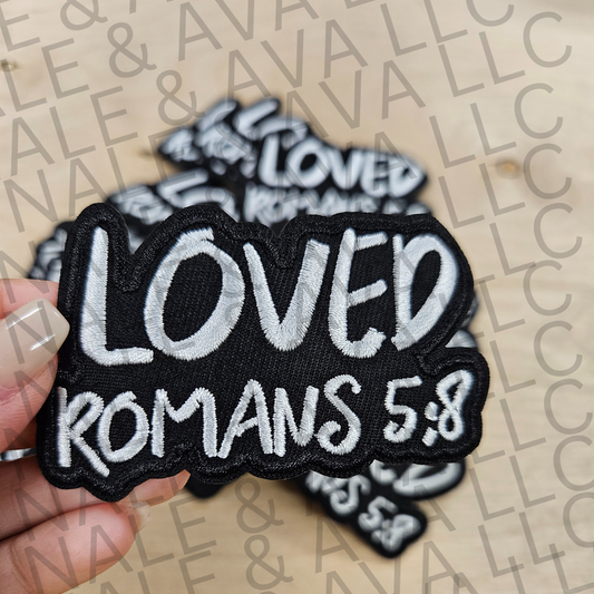 Black and white 'LOVED ROMANS 5:8' patch held by a hand on a beige background with 'Avalon' branding.